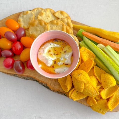 Wooden charcuterie board with vegetables, crackers, corn chips, and bowl of dip on white surface