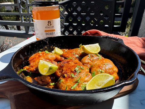 Chicken and lime wedges in black pan on patterned surface with jar of honey in background