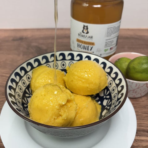 Bowl of mango sorbet in bowl on wooden surface with jar of honey and limes in the background