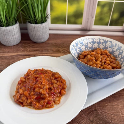 Baked beans on a white plate next to beans in a blue bowl, both on a white plate sitting on a wooden surface in front of green plants and a window.
