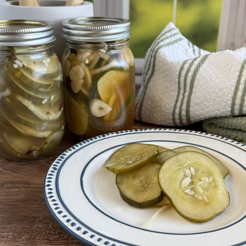Pickle slices on a white and blue plate in front of two jars of pickles and a white dish towel, all on a wooden surface.