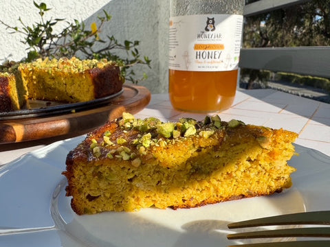 Slice of cake on white plate with honey jar and additional cake pieces in the background