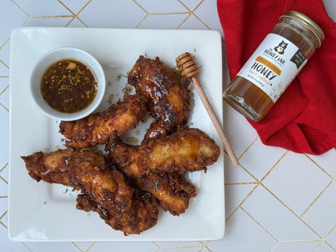 Chicken tenders on white plate with sauce and honey dipper. Jar of honey and red napkin sitting on patterned background on the side of the plate.