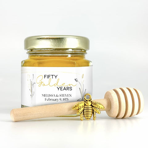 Small jar of honey with gold lid, gold bee charm, wooden dipper, and label with 'Fifty Golden Years' text on white background