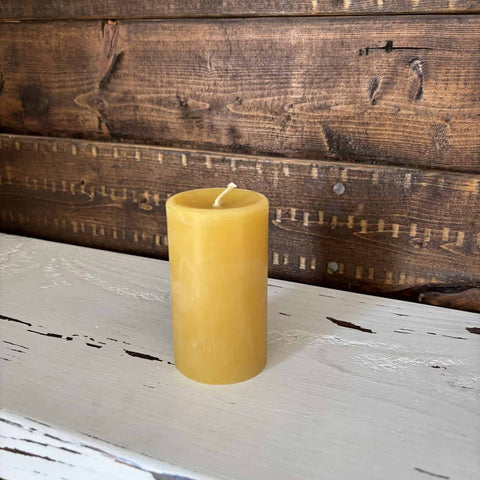 A single yellow beeswax pillar candle with a white wick sits on a distressed white wooden surface in front of a rustic dark brown wood plank wall.