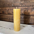 A single tall, cylindrical yellow beeswax candle with a cotton wick, standing upright on a distressed white wooden surface against a rustic dark brown wood plank background. 100% Beeswax Candle - 2" Wide Pillar Trio - The Honey Jar  