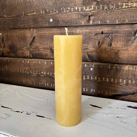A single tall, cylindrical yellow beeswax candle with a cotton wick, standing upright on a distressed white wooden surface against a rustic dark brown wood plank background. 100% Beeswax Candle - 2" Wide Pillar Trio - The Honey Jar  