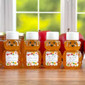 Four bear-shaped honey bottles filled with amber honey, arranged on a wooden table in front of a blurred kitchen window with red apples in the background. Each bottle has a white cap and a colorful custom label with names and Happy & Healthy New Year text.