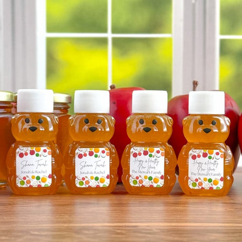 Four bear-shaped honey bottles filled with amber honey, arranged on a wooden table in front of a blurred kitchen window with red apples in the background. Each bottle has a white cap and a colorful custom label with names and Happy & Healthy New Year text.