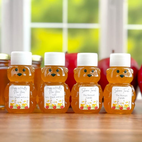 Four small bear-shaped honey jars with white lids and custom printed labels, arranged on a wooden table with red apples and a blurred window in the background.