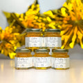 Five small jars with labels on a reflective surface with gold decorations in the background