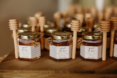 Small glass jars of honey with gold lids and white patterned labels, each paired with a wooden honey dipper on a rustic wooden surface.