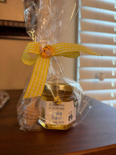 A honey jar wrapped in clear cellophane and tied with a yellow gingham ribbon, sitting on a wooden table in front of window blinds.