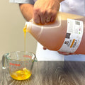 Person pouring honey from a large container into a Pyrex measuring cup on a wooden surface.