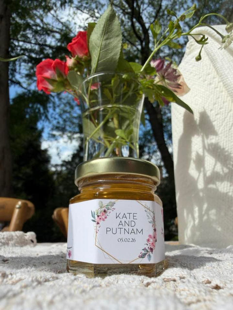 A small clear glass jar filled with honey, topped with a gold metal lid and a white floral label reading 'KATE AND PUTNAM' with a date. The jar is placed outdoors on a textured white surface, with a clear glass vase holding red flowers, against a blurred background of trees and sky.
