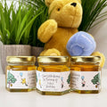 Close up of honey birthday party favor jars topped with gold lids. Jars have labels featuring classic Winnie the Pooh and Piglet holding blue balloons and a Our Little Hunny is Turning 4 title. Table decor and Winnie the Pooh plush toy in background.