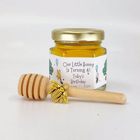 2 oz hexagon jar filled with honey, gold lid, gold bee charm, 3" wooden honey dipper and personalized Winnie the Pooh design on label with blue balloons and Our Little Hunny is Turning 4 title.