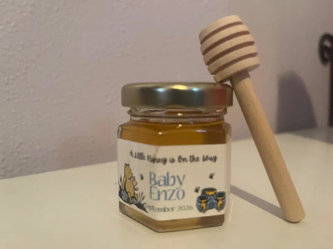 A small glass jar of honey with a gold lid and a custom white label reading 'Baby Enzo', with a wooden honey dipper resting against it on a white surface.