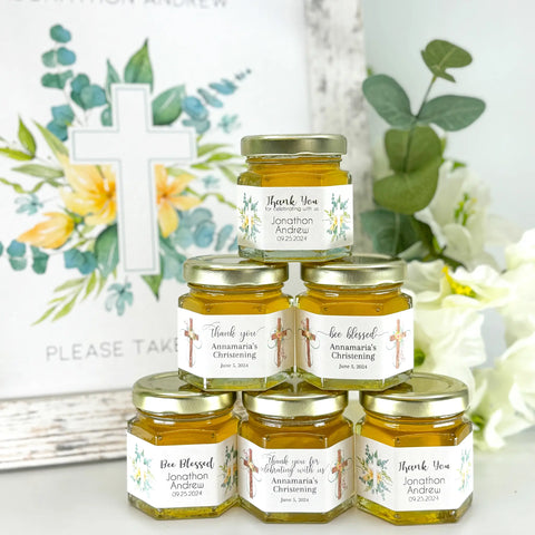 2 oz honey baptism favors on hexagon jars with gold lid with personalized floral swirls and crosses themed labels. Labels feature a variety of titles, including Thank you, Bee Blessed, and Thank you for celebrating with us. Free matching printable table sign in background.