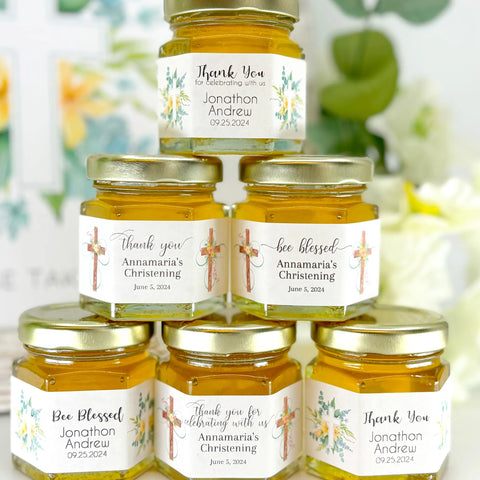 Close up of 2 oz honey baptism favors on hexagon jars with gold lid with personalized floral swirls and crosses themed labels. Shown label titles are Thank You, Bee Blessed, and Thank you for celebrating with us. Table decorations in background.