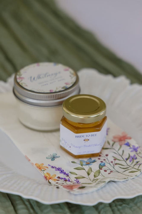 A small white jar with a silver lid and a hexagonal glass honey jar with a gold lid labeled Bride to Bee arranged on a floral paper napkin over a white scalloped plate with a green background.