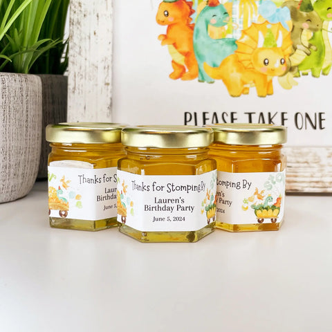 Close up of honey birthday party favor jars topped with gold lids. Jars have labels featuring dinosaurs with party balloons and hats and a Thanks for Stomping By title. Table decor in background.