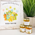 Party favor table decor is shown including a plant and honey birthday party favor jars topped with gold lids. Jars have labels featuring dinosaurs with party balloons and hats and a Thanks for Stomping By title. Matching free printable sign is also shown.