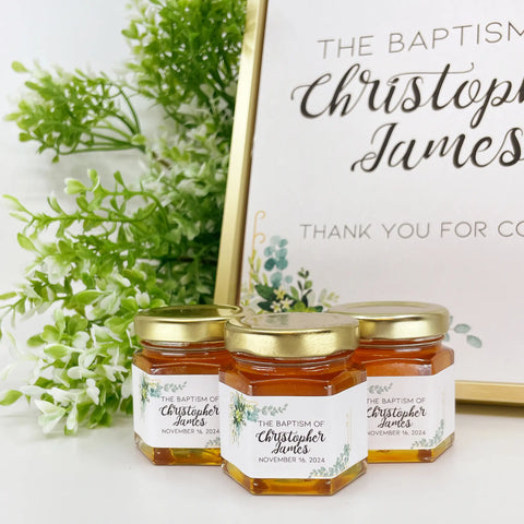 Group of 2 oz honey baptism favors in hexagon jars with gold lid with personalized labels. Labels feature gold frame with eucalyptus leaves. Label title shown is The Baptism of Christopher James. Table decorations in background.