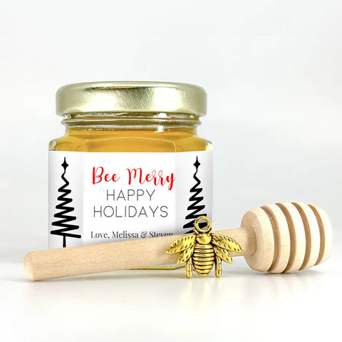 2 oz hexagon honey favor. Jar is filled with honey and has gold lid, 3" wooden honey dipper, gold bee charm, and personalized fine line trees label with title Bee Merry.