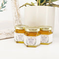 Close up of 2 oz honey baby shower favors in hexagon jars with gold lid and personalized geometric labels with the title Baby to Bee. Label features gold geometric frame with leaves. Table decorations in background.