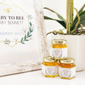 Baby Shower favor table decor shown with 2 oz hexagon favor jars filled with honey and topped with gold lid. Jars have label featuring geometric design and Baby to Bee title. Table shows free printable sign and plant in background.