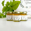 Close up of 2 oz honey thank you wedding party shower favors on hexagon jars with gold lid with personalized geometric labels. Labels feature gold geometric frame with leaves. Label title is Thank You. Table decorations in background.