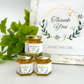 2 oz honey thank you wedding party shower favors on hexagon jars with gold lid with personalized geometric labels. Labels feature gold geometric frame with leaves with Thank You title. Free matching printable table sign in background.