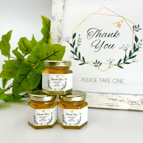 2 oz honey thank you wedding party shower favors on hexagon jars with gold lid with personalized geometric labels. Labels feature gold geometric frame with leaves with Thank You title. Free matching printable table sign in background.