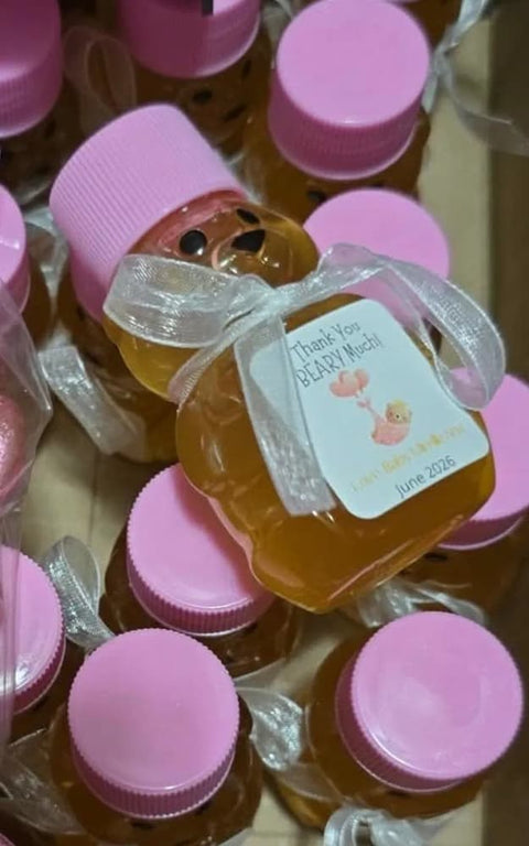 A close-up of several small honey jars with pink screw-top lids, arranged together. The central jar has a white ribbon bow with a printed label that reads 'Thank You Beary Much!'.