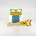 2 oz hexagon jar filled with honey, gold lid, 3" wooden honey dipper, gold bee charm, and personalized blue graphics design on label.