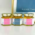 Group of 2 oz honey baptism favors in hexagon jars with gold lid with personalized labels. Labels feature pink or blue graphics-style design. Table decorations in background.