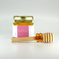 2 oz hexagon jar filled with honey, gold lid, 3" wooden honey dipper, emanel honeycomb charm, and personalized pink graphics design on label.