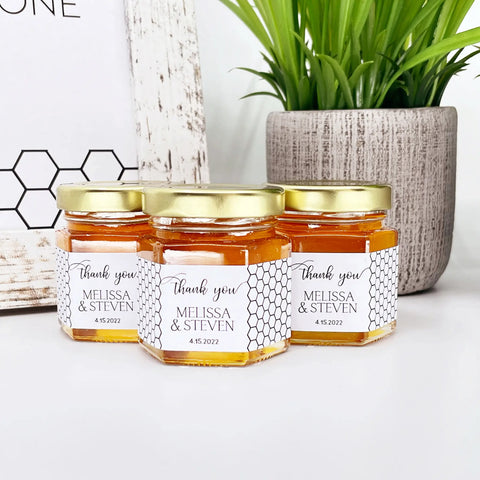 Close up of 2 oz honey thank you wedding party shower favors on hexagon jars with gold lid with personalized honeycomb labels. Labels feature black and white honeycomb design and Thank You title. Table decorations in background.