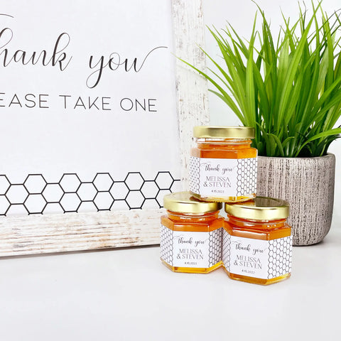 2 oz honey thank you wedding party shower favors on hexagon jars with gold lid with personalized labels. Labels feature black and white honeycomb design with Thank You title. Free matching printable table sign in background.