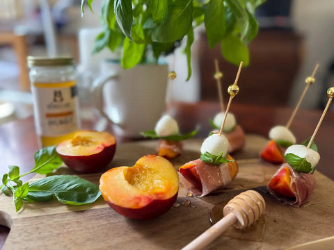 Wooden board with prosciutto-wrapped peaches, mozzarella balls, and basil leaves, accompanied by a jar of honey.