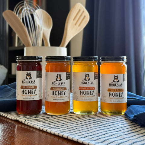 Four glass jars of Honey Jar honey arranged on a striped placemat over a wooden table, with kitchen utensils and a dark curtain blurred in the background. The jars contain different honey shades: dark red, amber, golden yellow, and orange.