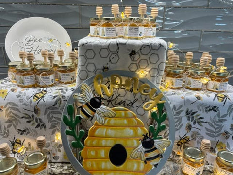 A styled honey-themed display featuring multiple small glass jars of honey with wooden dippers, a white plate reading 'Bee Happy,' patterned table linens, and a round decorative sign in front with a beehive, bees, and the word 'honey'.