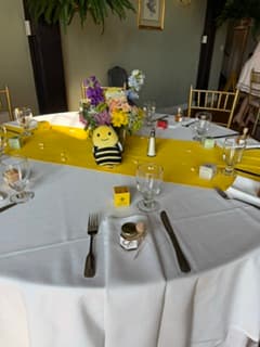 A round dining table set with a white tablecloth, silver cutlery, clear drinking glasses, small jars of honey, and a bright yellow table runner. A floral centerpiece and a small yellow-and-black bee decoration sit in the center of the table.