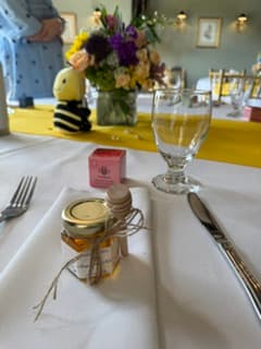 A small glass jar filled with honey, tied with twine and set on a white tablecloth at a dining table. A glass, knife, fork, flowers, and a yellow table runner are visible in the background.