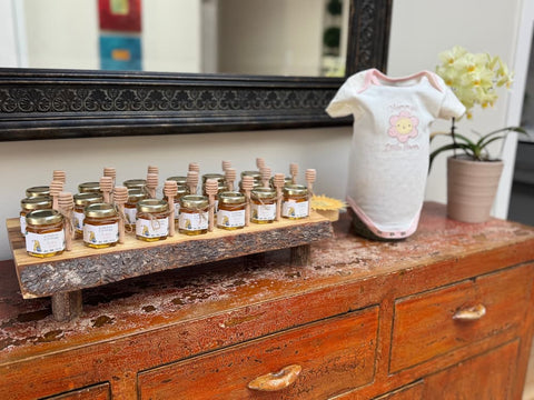 A set of small glass jars filled with honey, each fitted with a wooden honey dipper, displayed on a rustic wooden stand on top of a distressed brown wooden dresser. A white baby onesie with a pink flower graphic stands next to them, and a potted yellow orchid is visible in the background.