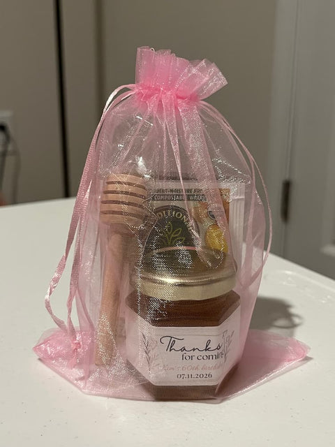 A honey gift set wrapped in a sheer pink organza bag, containing a jar of honey, a wooden honey dipper, and bags of tea.