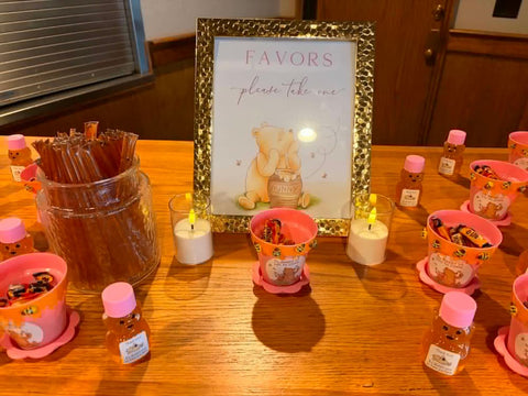 A wooden table set up as a favor station with a central framed sign reading 'FAVORS please take one' above a teddy bear illustration. Several pink cups, small honey bottles with pink lids, a glass jar of wooden straws, and two lit white candles are arranged around it.