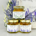 Close up of 2 oz honey thank you wedding party shower favors on hexagon jars with gold lid and personalized labels. Labels feature two lavender designs. Label titles shown are Thank You, Thanks for Bee-ing Here, and Thanks for Coming. Lavender florals in background.