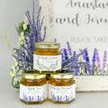 2 oz honey thank you wedding party shower favors on hexagon jars with gold lid and personalized labels. Labels feature two lavender designs with a variety of titles. Title shown include Thank You, Thanks for Bee-ing Here, and Thanks for Coming. Matching printable table sign and lavender florals shown in background.